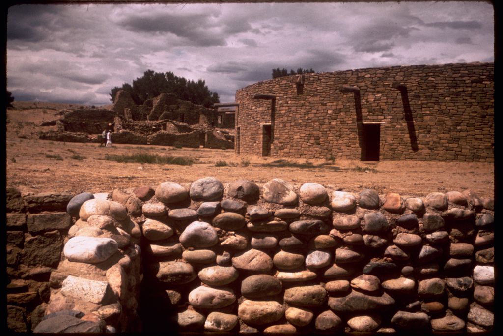A low wall of large rounded stones stands before taller coursed masonry rooms under a cloudy sky, with a dark slide border around the image.