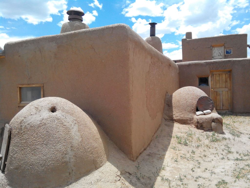 Two rounded earthen ovens stand beside thick brown plastered walls with wooden doors, small windows, roof drains and modern vents.