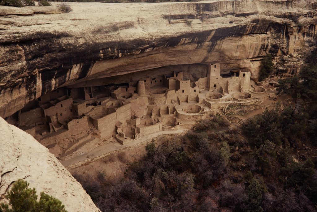 A large complex of stone rooms, round structures and towers occupies a deep rock alcove beneath a dark overhang.