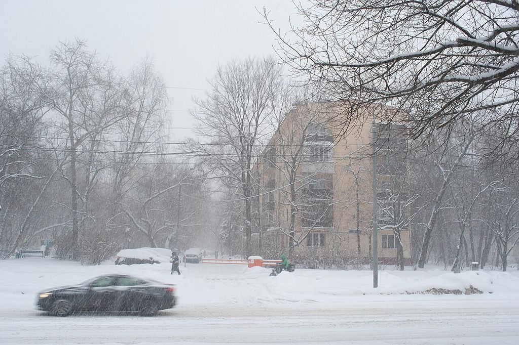 A tan five-storey apartment block is partly hidden by bare trees and falling snow, with pedestrians, parked cars and a blurred car on the foreground road.