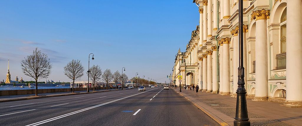 A broad riverside road recedes beside long pale palace façades, with the Neva and a distant gilded spire across the open left side.
