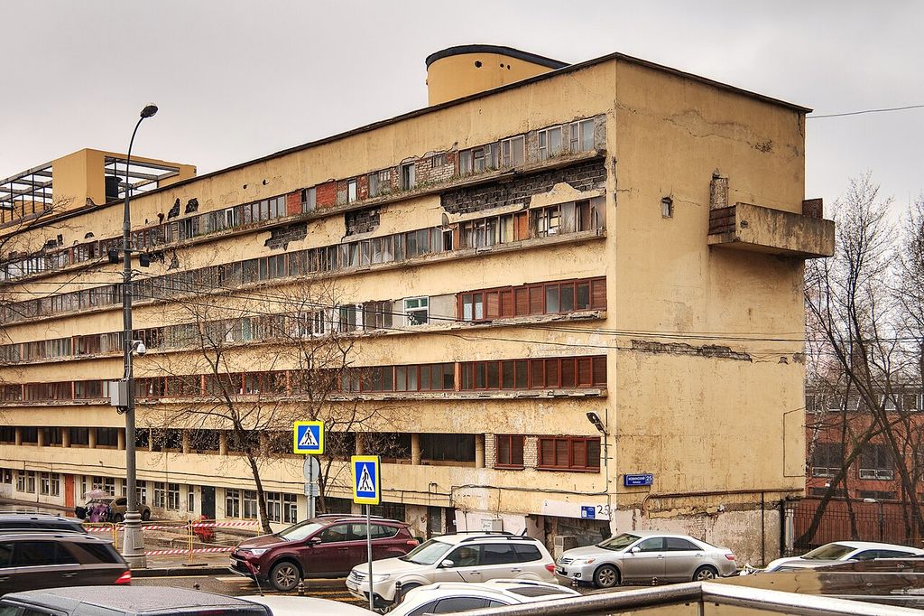 A long weathered cream apartment block with horizontal window bands, altered glazing, cracked render and a projecting end balcony stands above a busy road.