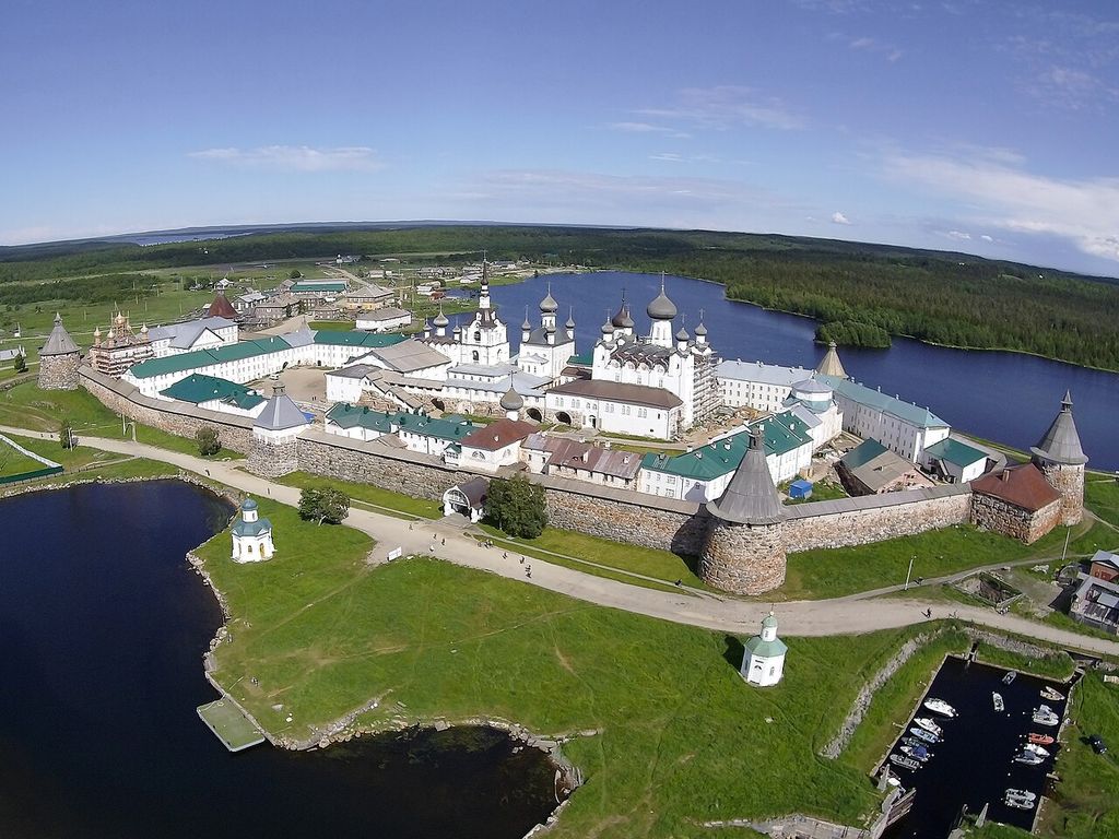 An aerial view shows a large walled monastery with round stone towers, white churches, long ranges, roads and yards between dark blue water and forest.