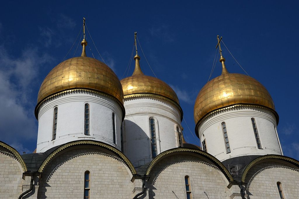 Three large gilded domes rise on white cylindrical drums above curved gables and narrow windows against a deep blue sky.