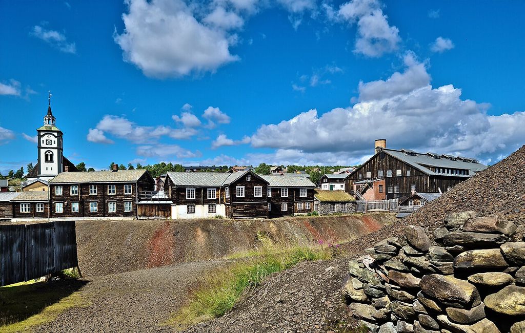 Dark timber houses and a white clock-tower church stand behind broad grey slag heaps, with a large timber smelter building at right under a bright blue sky.