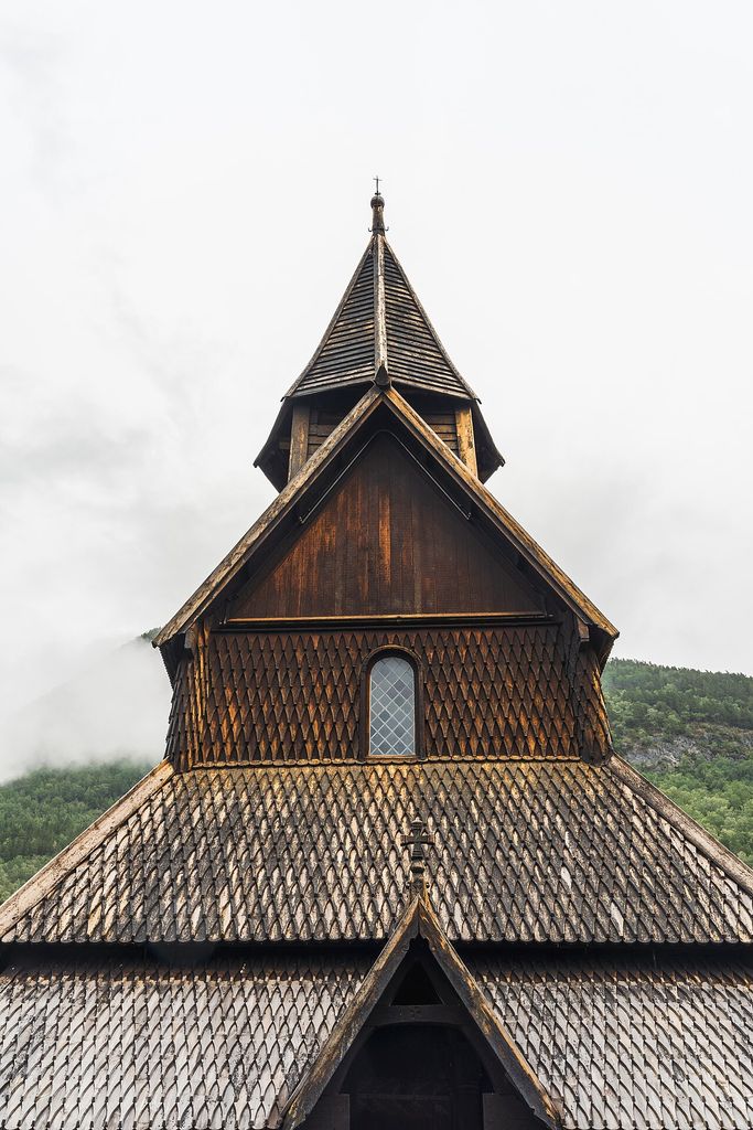 A close frontal view of a dark timber stave church shows steep overlapping shingle roofs, triangular gables, a small window and a narrow turret.