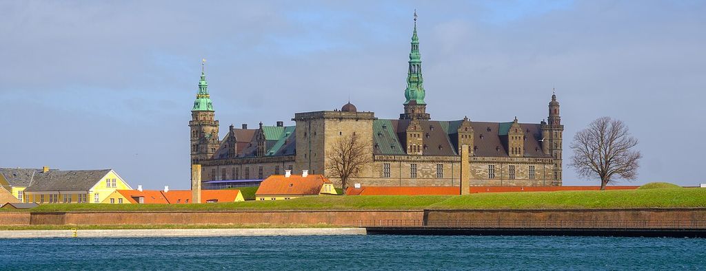 A long Renaissance castle with stone towers, copper roofs and a central spire stands behind low grassy ramparts across blue harbour water.