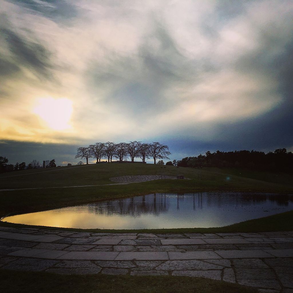 Stone paving leads past a reflecting pond toward a low grassy hill crowned by a small grove of bare trees beneath a dramatic evening sky.