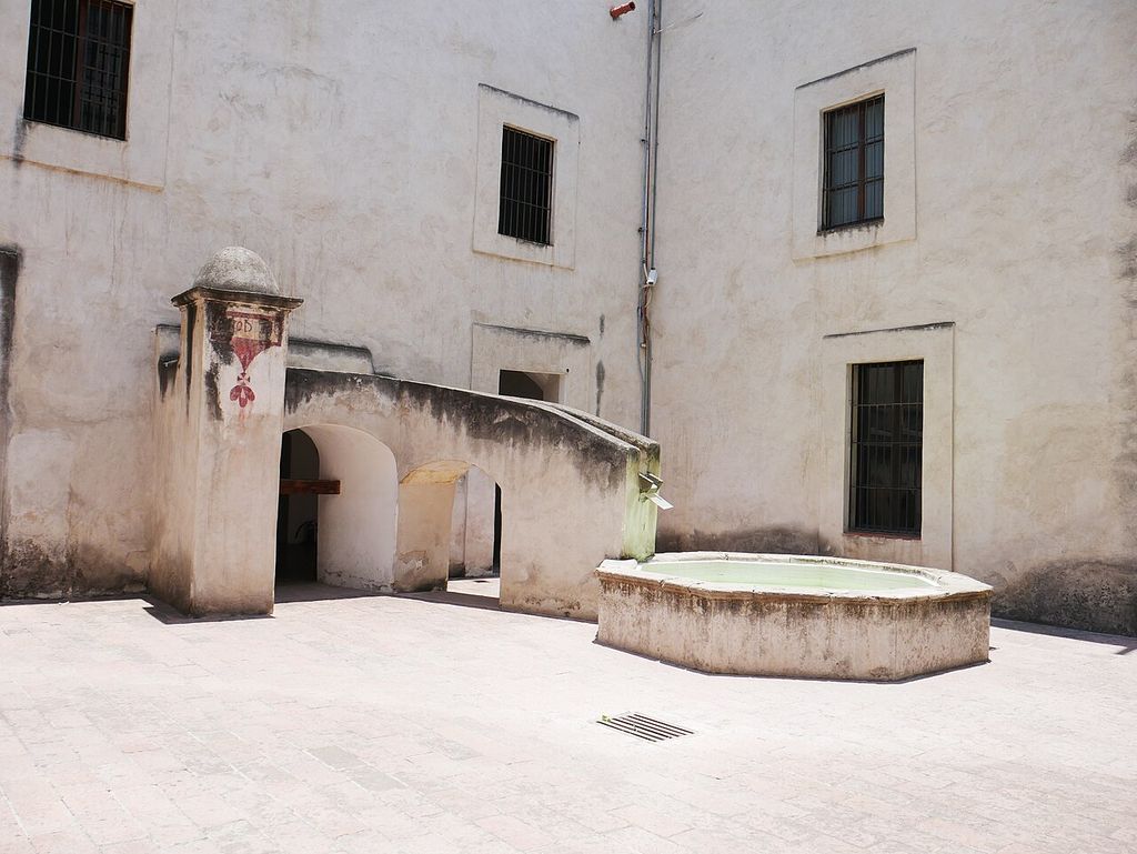A low basin stands in a paved court beside plastered walls, barred windows and a raised stair with arched access.