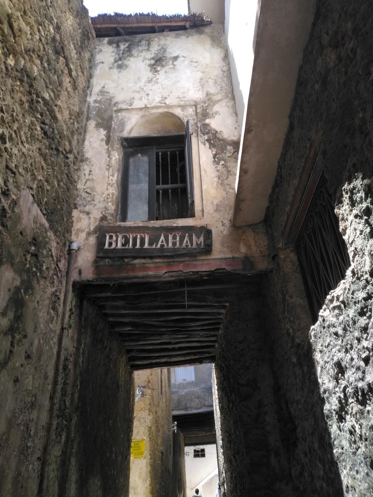 A timber bridge links the upper floors of weathered stone buildings above an extremely narrow, shadowed lane.