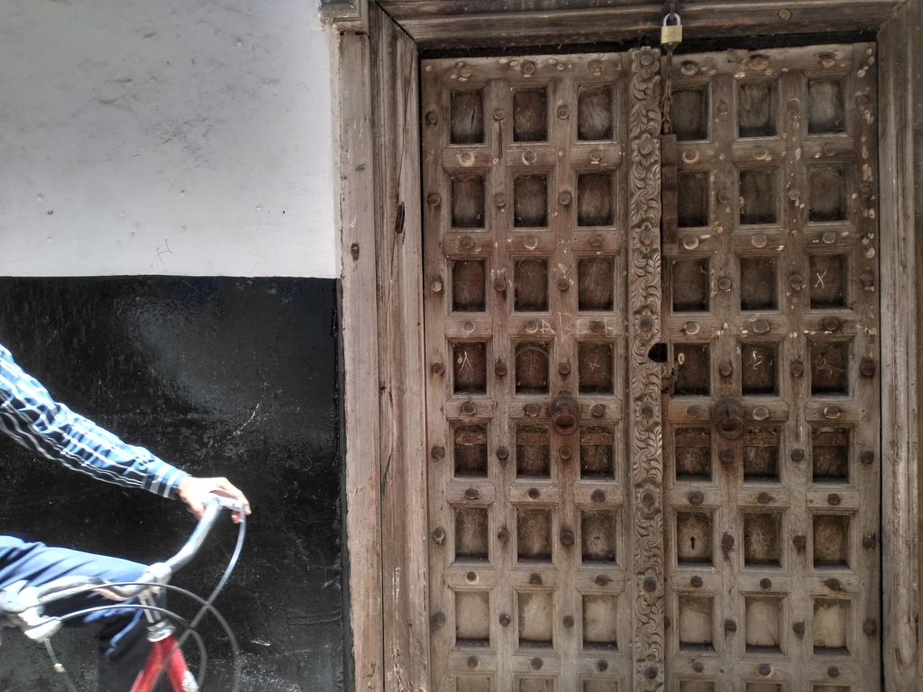 A large double timber door has deep square panels, rows of metal bosses, ring handles and a carved central strip, with part of a passing cyclist at left.