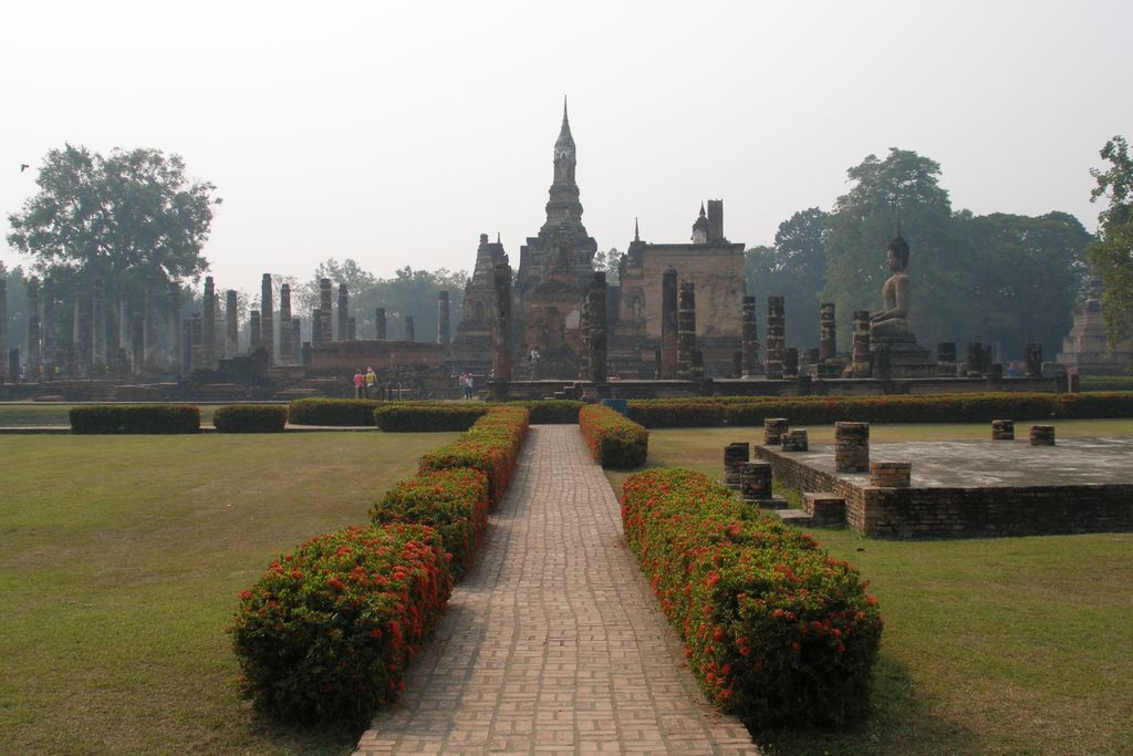 A brick path crosses lawns toward weathered temple towers, columns and Buddha images in a hazy archaeological park.