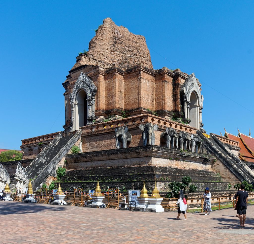 A massive partly ruined brick chedi with carved stair guardians rises above a paved court and visitors under blue sky.