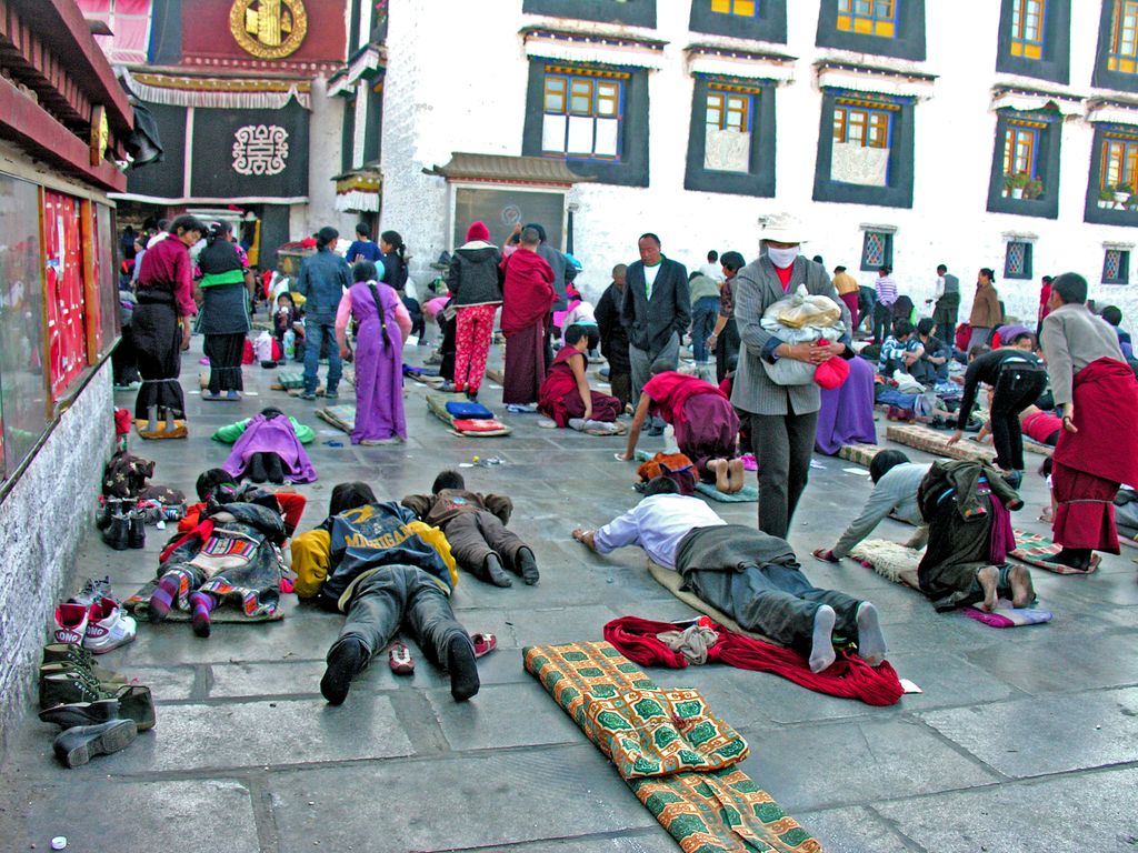 Pilgrims lie full-length or kneel on a stone court before a colourful temple entrance while other worshippers gather nearby.