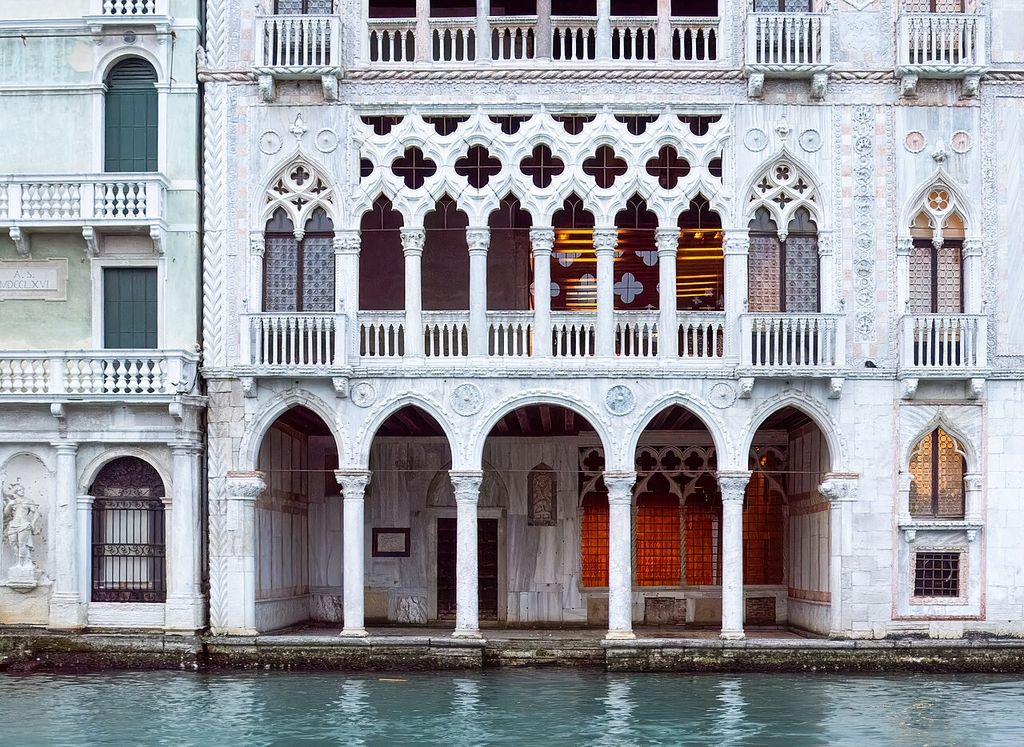 A close canal view shows a pale stone palace with an open water-level arcade, two stacked loggias, pointed arches, quatrefoils, balconies and narrow side windows.