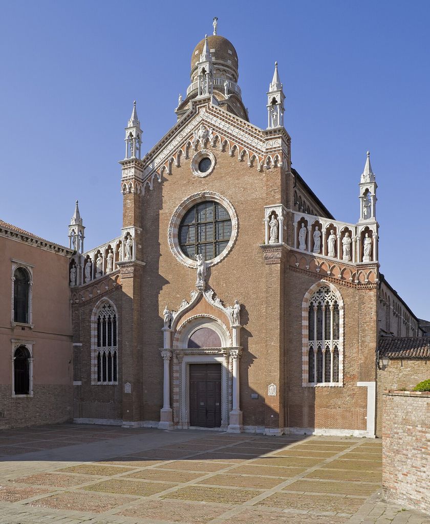 A tall red-brick church façade has a large circular window, pointed side windows, a carved pale stone portal, rows of statues and small corner pinnacles.