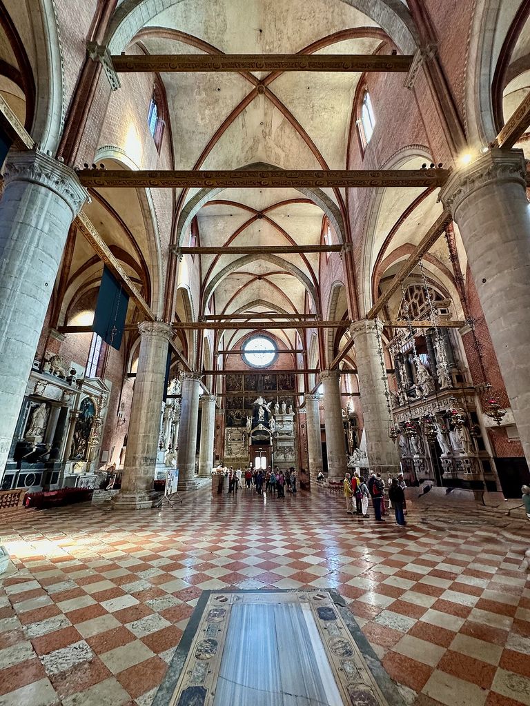 A tall church interior has red brick walls and vault ribs, pale cylindrical piers, pointed arches, timber tie beams, a patterned tile floor and many later monuments.