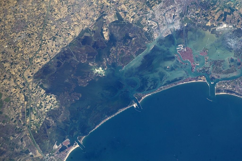 An orbital view shows the compact reddish islands of Venice within a broad green-blue lagoon, separated from the dark Adriatic by a long barrier beach and surrounded by marshes and mainland fields.