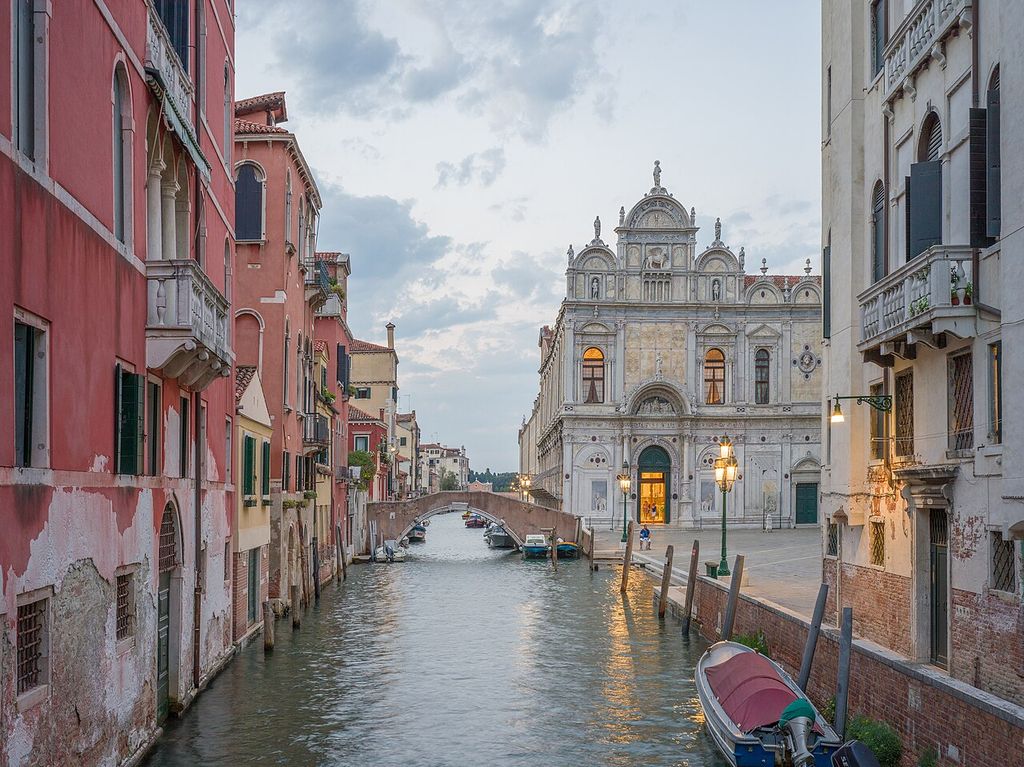 A narrow canal lined by red and white houses leads toward an ornate pale stone civic façade, with a small bridge, boats and lit entrances at dusk.