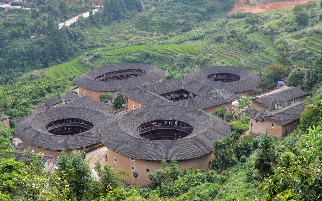 Circular and oval earthen buildings surround a square building, each enclosing an open court amid cultivated hills.