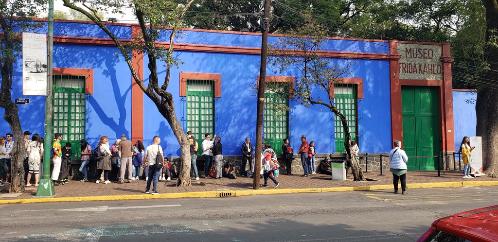 Blue exterior walls and green windows of Casa Azul, with trees and visitors along the pavement.