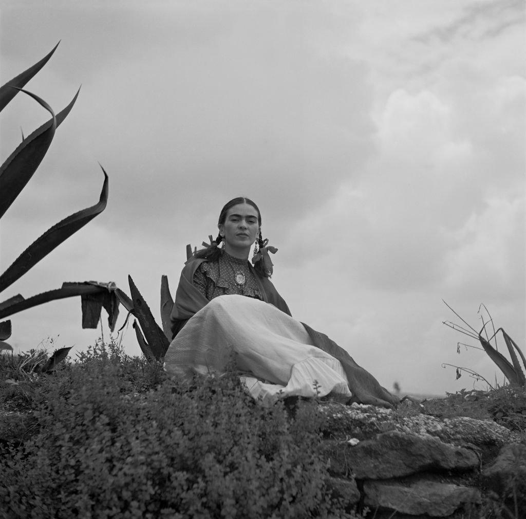 Frida Kahlo seated outdoors beside the pointed leaves of an agave, against an open sky.