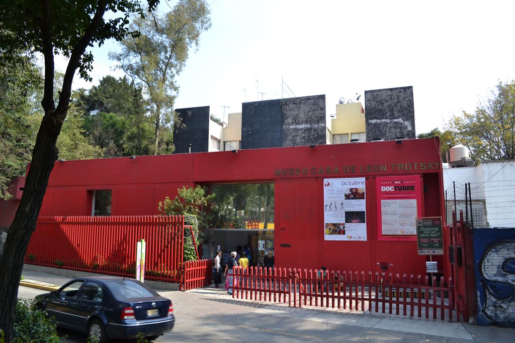 Red street entrance to the Trotsky museum, behind a red fence and trees.