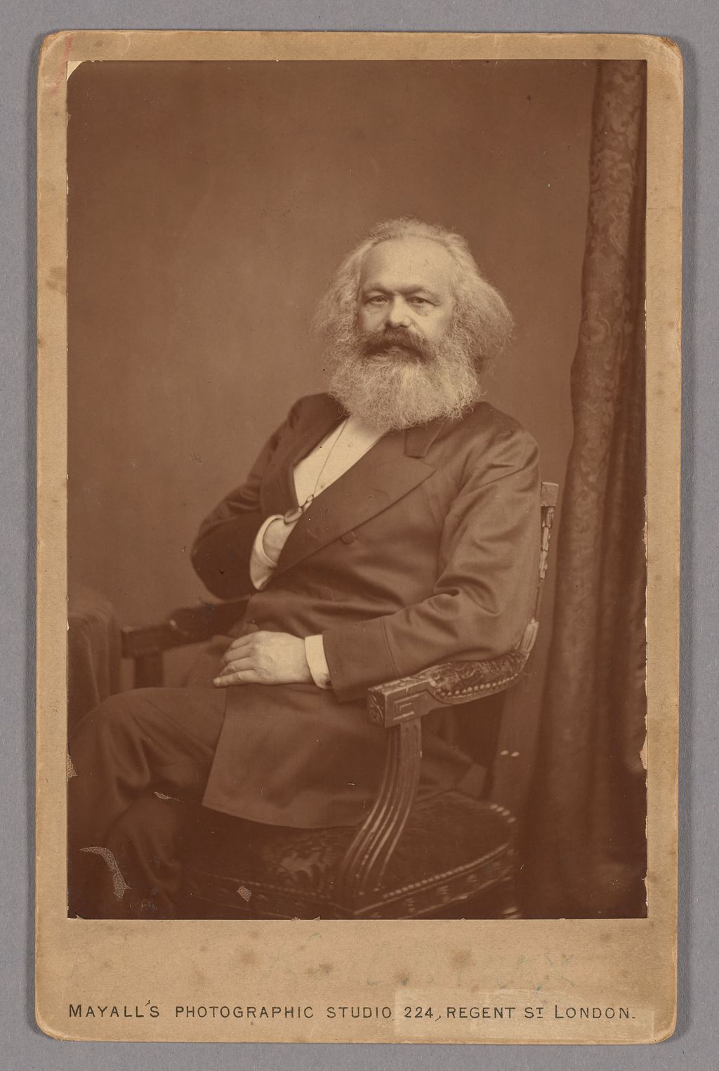 Historical portrait of Karl Marx with a white beard, seated in a dark suit.