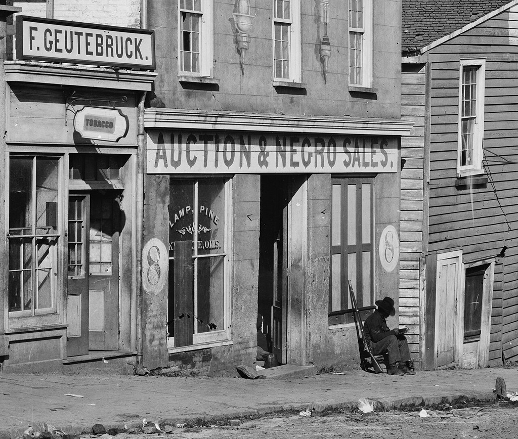 Nineteenth-century building with a slave-auction sign and an unidentified person seated beside it.