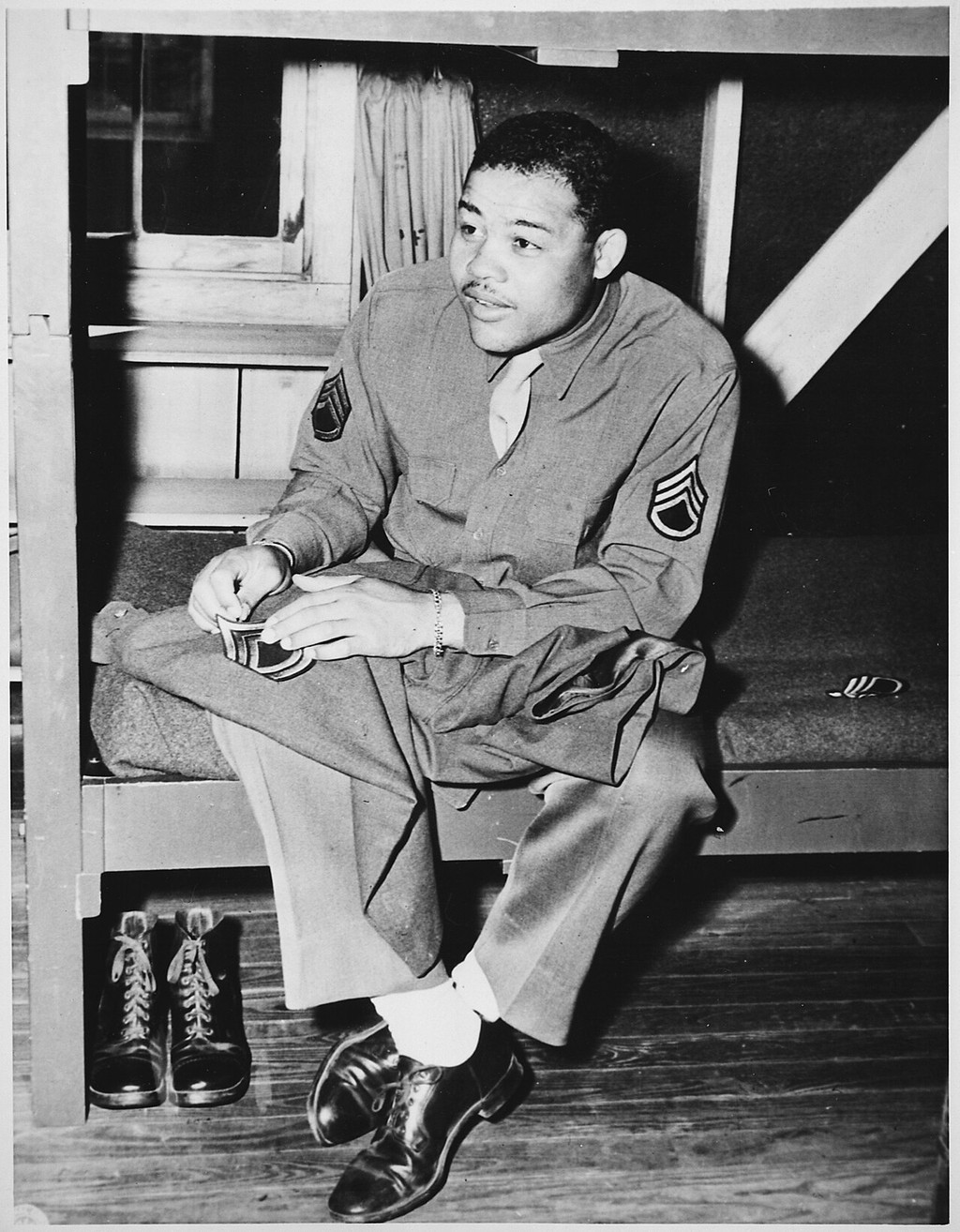 Joe Louis seated in Army uniform, sewing stripes onto a garment on his lap.