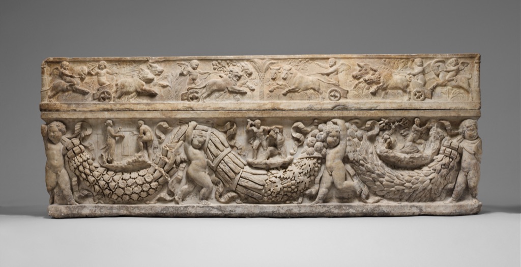 The Roman marble sarcophagus has repeated hanging garlands and framed relief scenes arranged across a long carved box and lid.