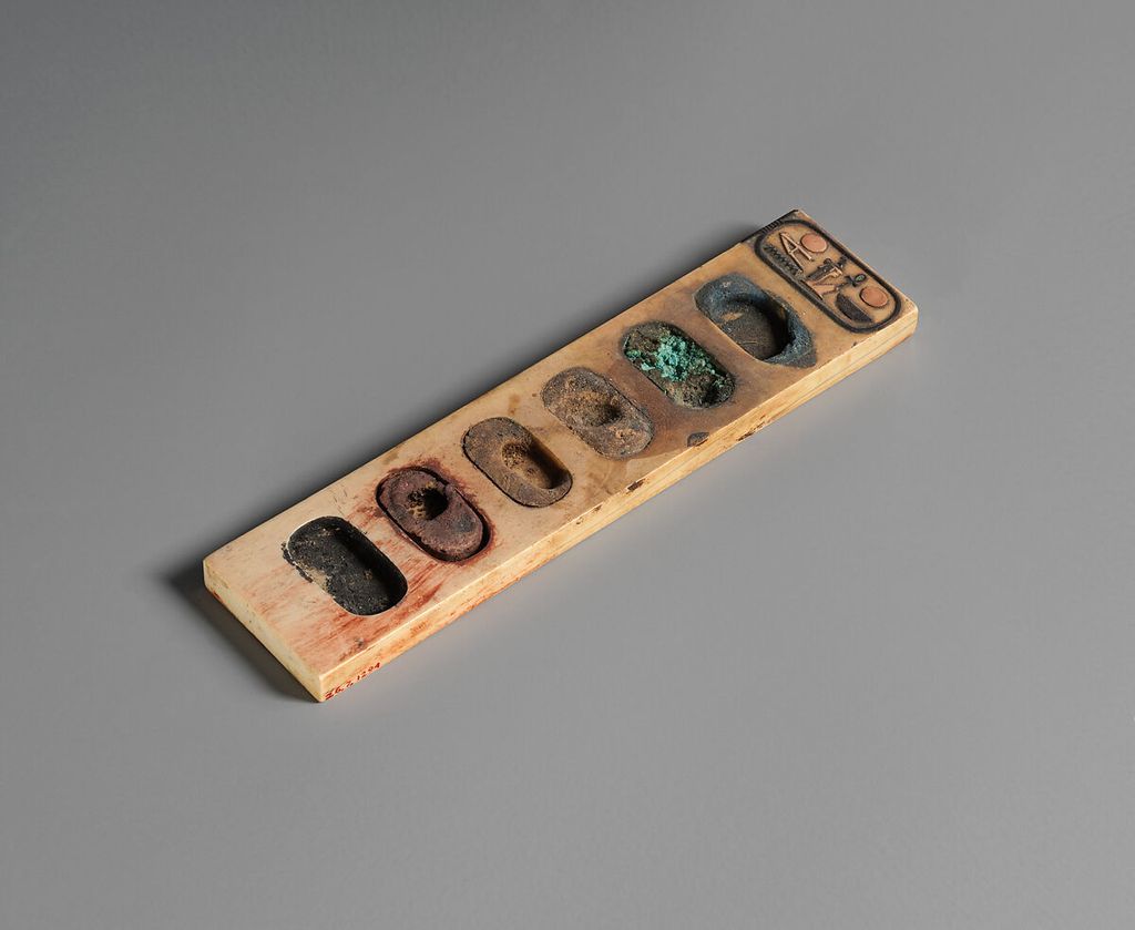An elongated ivory painter’s palette has six oval wells retaining cakes of blue, green, brown, yellow, red and black pigment.