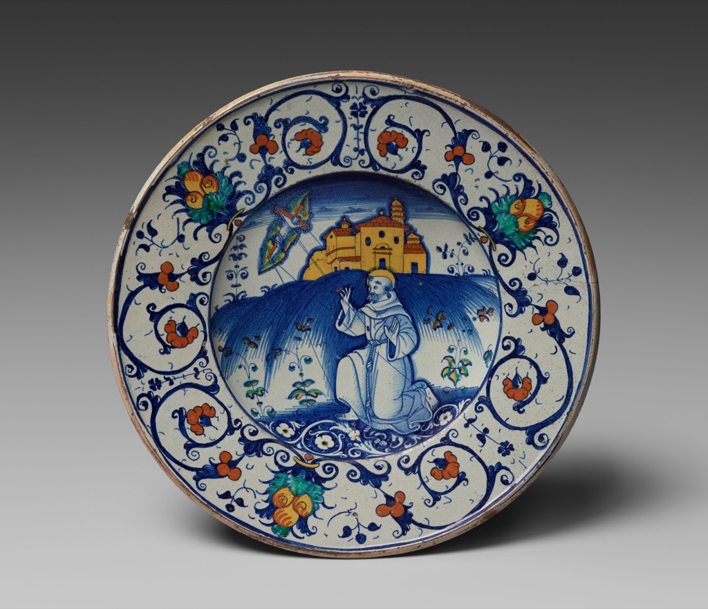 A circular pale-ground maiolica bowl uses dense blue drawing with orange-red flowers, yellow-orange architecture and small green leaves.