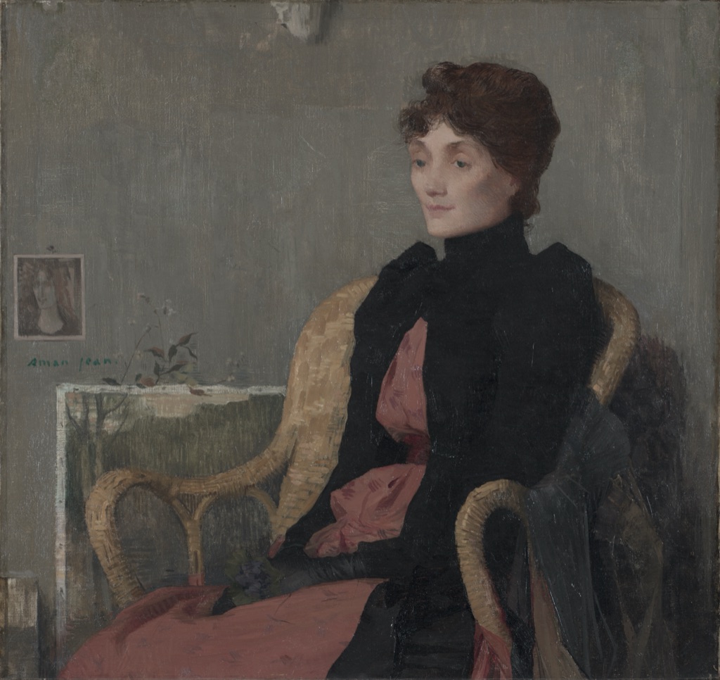 A seated woman in a black jacket and muted pink dress appears against a restrained gray-green interior with wicker chair and small wall picture.