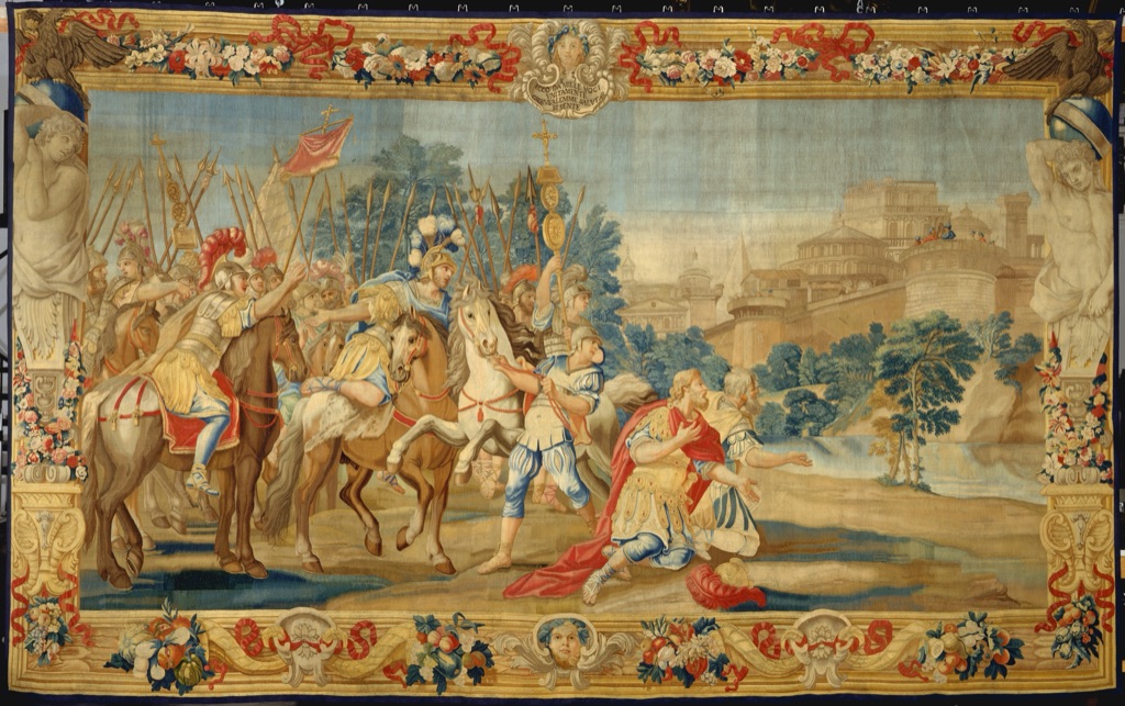 A wide tapestry shows mounted and kneeling figures in red, blue and yellow garments before pale city walls within a flowered border.