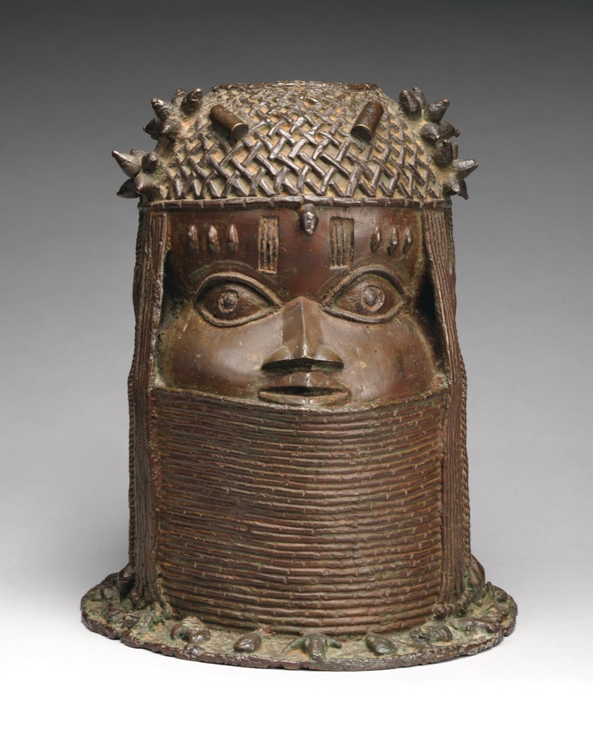 A brass commemorative head of an oba has a tall patterned crown, projecting facial features and a cylindrical neck.