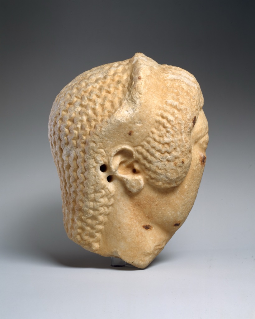 A pale marble woman's head turns slightly with broad cheek planes, carved eyes and patterned hair.