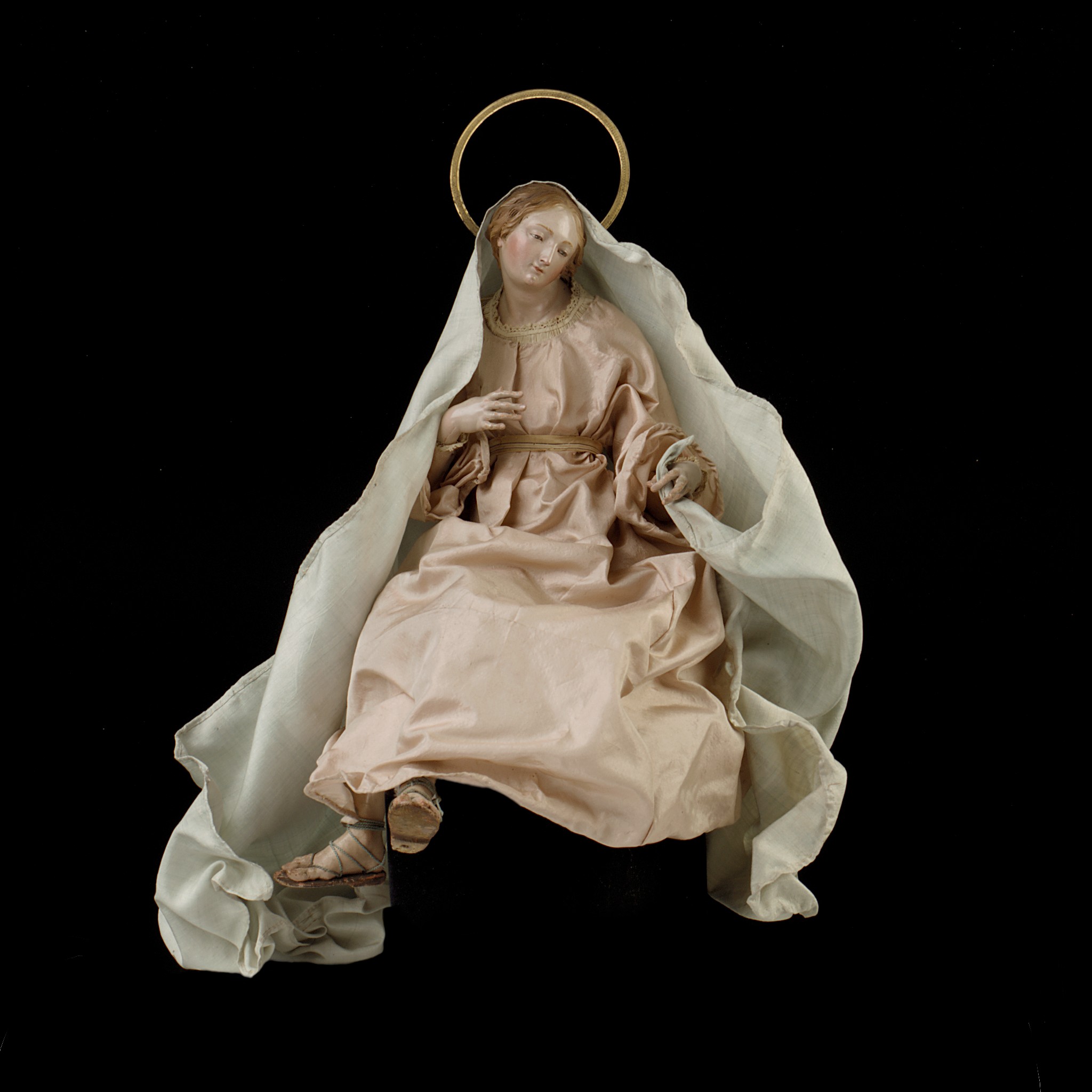 Seated crèche Virgin with a terracotta head, articulated hands, pale garments and a circular halo over a wire-and-tow body.